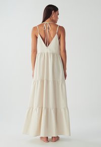 Moteris vilki be rankovių, sluoksniuotą beigi maksi suknelę su gilia V formos nugara, surišta ant kaklo, stovinti atsukusi nugarą ant vienodo fono.