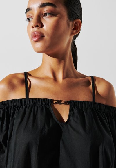 Haut noir décolleté avec des bretelles réglables, encolure froncée, accent en forme de trou de serrure et détail en matériel brillant au centre. Texture lisse.