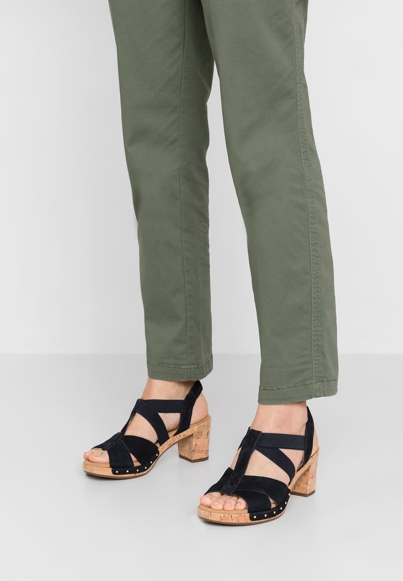 Personne portant un pantalon vert olive et des sandales à talons blocs en liège noires à lanières, debout sur un sol gris clair.