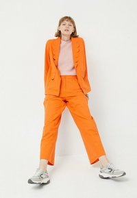Blazer naranja a medida con detalles en forma de ola, pantalones naranja a juego y zapatillas grises. Top texturizado rosa usado debajo.