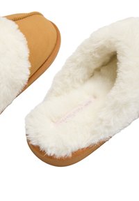 Zapatilla color canela con un suave forro de piel sintética blanca, punta redondeada y suela texturizada. Presenta un diseño cosido a lo largo del borde.