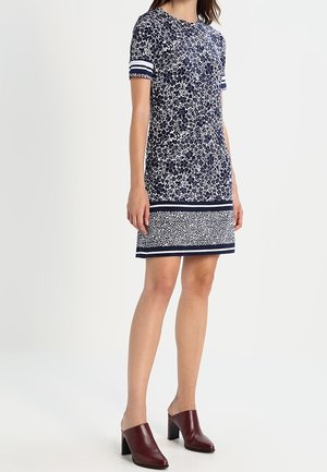 Mujer con vestido de manga corta con estampado floral en azul marino y blanco, con puños y dobladillo a rayas, combinado con mules de tacón de color rojo oscuro.