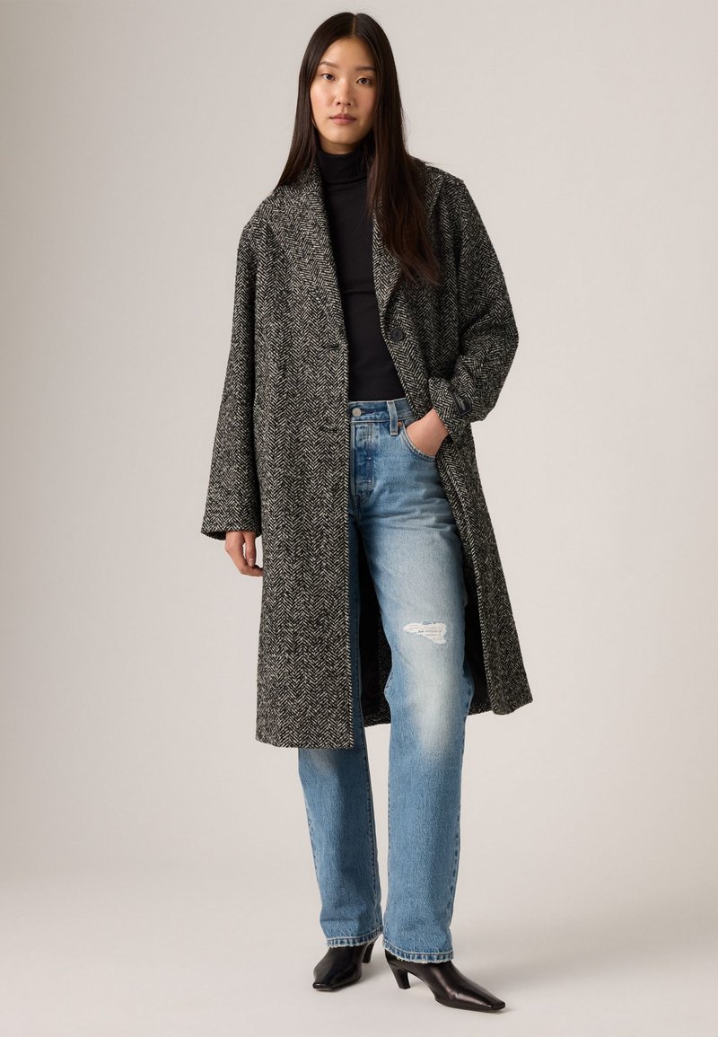 Langer, grauer Fischgrätmantel mit lässigem Schnitt, getragen über einem schwarzen Rollkragenpullover, kombiniert mit hellblauen, verwaschenen Jeans und schwarzen Stiefeletten mit Absatz.