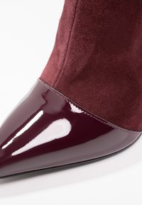 Bottine bordeaux à bout pointu, avec un bout en cuir verni brillant et une tige en daim mat, soulignée par des détails de coutures.