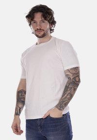 T-shirt bianca in cotone con maniche corte, scollo rotondo e vestibilità casual, indossata da un modello con tatuaggi, su uno sfondo bianco semplice.