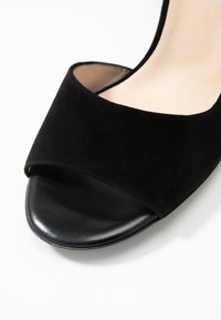 Talons ouverts en suede noir avec une semelle lisse en cuir noir et une large sangle avant pour un soutien. Design minimaliste, sans quincaillerie visible.