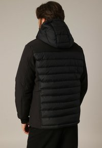 Homme de dos portant une veste matelassée noire avec une capuche, montrant un dos rembourré et des manches en tissu solide.