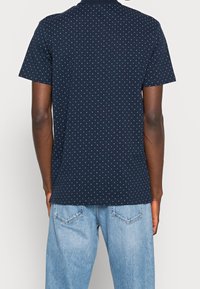 Námořnická modrá polo košile s krátkým rukávem, která má malý bílý puntíkový vzor a klasický límec. Doplňuje ji světlé modré džíny.