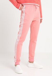 Rosa sweatpants har vita sidosträck, ett elastiskt midjeband med dragsko och en figurnära bendesign. Åtsidan med vita slip-on skor.