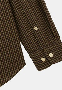 Chemise à manches checkée brune et noire, dotée de deux boutons beige clair, avec des coutures visibles et une poche poitrine.