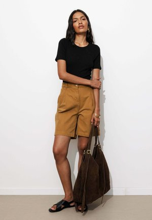 Femme debout contre un mur blanc, portant un haut noir à manches courtes, un short beige, des sandales noires et tenant un grand sac cabas en daim marron.