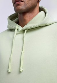 Lichtgroene hoodie van zacht materiaal, met een trekkoord in de capuchon en een ontspannen pasvorm. Getextureerd materiaal met een gladde afwerking.