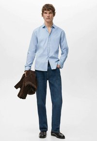 Camicia a righe blu e bianche con bottoni, indossata con jeans blu, che tiene una giacca marrone, scarpe nere, in piedi contro uno sfondo semplice.