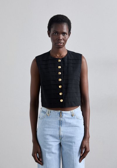 Black cropped sleeveless top with horizontal pleats and gold button detailing. Paired with light blue high-waisted jeans and a natural texture.