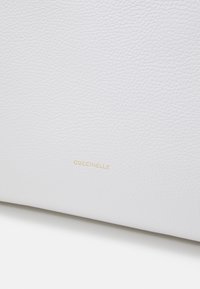 Surface en cuir blanc texturé avec le nom de la marque embossé en or "COCCINELLE" centré près du bas.