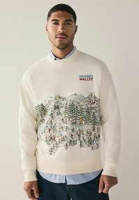 Cream sweatshirt featuring "Where's Wally?" graphic with colorful illustration of a wintry scene, trees, and numerous characters. Ribbed cuffs.
