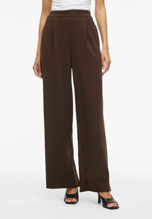 Femme portant un pantalon marron foncé taille haute à jambes larges, associé à des sandales noires à talons ouvertes sur un fond blanc.