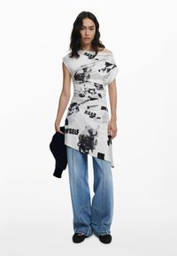 Weißes asymmetrisches Oberteil mit grafischen schwarzen Drucken, kombiniert mit hellblauen weit geschnittenen Jeans. Weiche Textur und entspanntes Design.