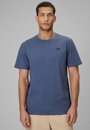 Mężczyzna w gładkim niebieskim t-shircie z krótkim rękawem i małym czarnym logo "4F" po lewej stronie klatki piersiowej, stojący na jednolitym tle.