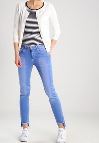 Valkoinen neuletakki, tummansininen raidallinen paita, vaaleansiniset kapealahkeiset farkut, joissa on rispaantuneet reunat, ja valkoiset tennarit. Pehmeä materiaali, rento muotoilu.