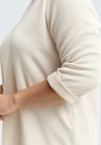 Vista laterale di una persona che indossa un top a maniche lunghe color crema a coste, con le maniche arrotolate fino all'avambraccio, su uno sfondo semplice.