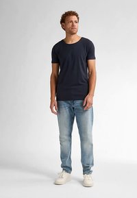 Navyblå kortærmet t-shirt med rund halsudskæring, båret med lyseblå jeans og hvide sneakers. Modellen står barfodet.
