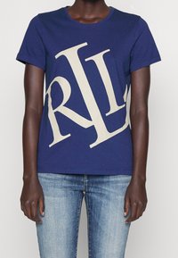 Osoba na sobě má tmavě modré tričko s velkými nakloněnými bílými písmeny "RL" a světle modré vybledlé džíny na jednotném pozadí.