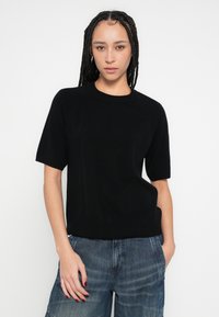 Μαύρο πλεκτό πουλόβερ με κοντά μανίκια, στρογγυλή λαιμόκοψη, ριμπ μανσέτες και τελείωμα, φορεμένο με τζιν παντελόνι άνετης εφαρμογής. Μαλακή υφή, casual εφαρμογή.