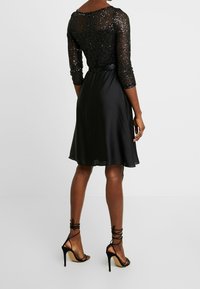 Robe noire avec un haut en sequins et une jupe en satin lisse. Le design comprend des manches trois-quarts et une taille cintrée par un lien.