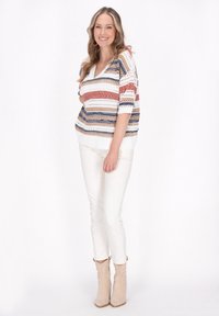 Maglione bianco lavorato a maglia con righe orizzontali in rosso, blu e oro, scollo a V e maniche corte, abbinato a pantaloni bianchi e stivali beige.