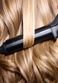 Schwarzes Haarstylingwerkzeug mit glatter Oberfläche, platziert auf welligem, hellblondem Haar. Das Werkzeug hat ein elegantes Design und eine Markenkennzeichnung.