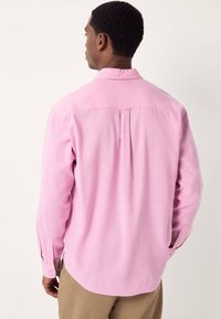 Vaaleanpunaisesta kankaasta valmistettu pitkähihaisen paidan, jossa on nappikiinnitteinen kaulus, takaosassa poimutus ja nappikiinnitteiset kalvosimet. Helma on kaareva.