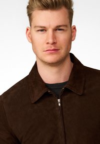 Brown suede jacket with a zipper, featuring a collar and smooth texture. Worn over a dark knitted sweater. Neutral background.