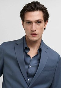 Blauer Blazer über einem Hemd, das ein elegantes Design, glattes Material und eine einzelne Brusttasche aufweist. Die Textur wirkt maßgeschneidert und passgenau.