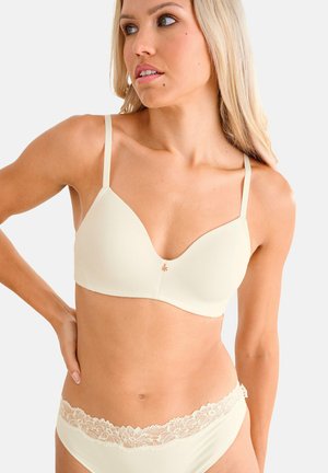 Vrouw met lang blond haar draagt een crème-achtige bh en bijpassend ondergoed met kanten rand, kijkt opzij met hand op heup.