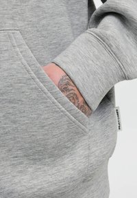 Grå hoodie med kengurulomme. Stoffet har en blød tekstur med en blanding af lyse og mørke grå nuancer. Mærket læser "PROJECT CORE."