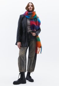 Sciarpa di lana colorata con lunghe frange, sovrapposta a un top grigio e un blazer di pelle nera. Indossata con jeans chiari strappati e stivali combat neri.