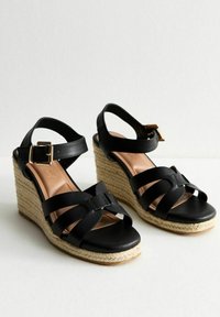 Black wedge sandals with wide cross straps, a buckled ankle strap, and a woven jute heel. Smooth leather upper and cushioned insole.