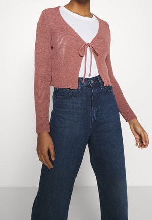 Vrouw draagt een kort, stofroze gebreid vestje dat op de borst is gestrikt, over een wit T-shirt en een donkerblauwe jeans met hoge taille.