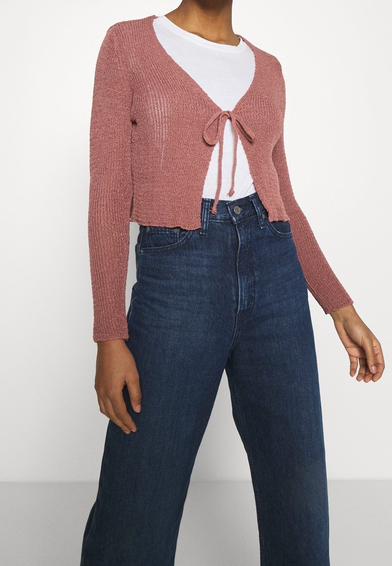 Femme portant un cardigan tricoté court rose poudré noué au niveau de la poitrine, sur un T-shirt blanc et un jean taille haute bleu marine.