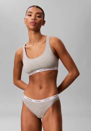 Femme portant un soutien-gorge gris clair de la marque Calvin Klein et une lingerie assortie sur fond gris uni, les mains dans le dos.
