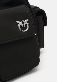 Saco de tecido preto com bolso frontal e fecho de argola prateada, apresentando dois pássaros a olhar um para o outro sobre um fundo branco.