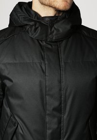 Chaqueta acolchada negra con cuello alto, cierres de presión y bolsillos laterales. Textura lisa con detalles acolchados en los hombros.