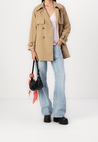Beige trenchcoat met brede revers en dubbel knoopsgat, gecombineerd met lichtblauwe flared jeans en een zwarte tas versierd met een rode patroon sjaal.