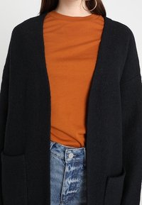 Musta, ylisuur neulejakku pehmeää kangasta, avoimella edustalla, kaksi etutaskua, yllään oranssi pyöreäkauluksinen paita ja vaaleansiniset farkut.