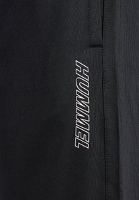 Sort stof med vertikal hvid "HUMMEL"-logo trykt langs sømmen på sportsbeklædning.