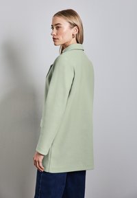 Cappotto verde chiaro con un design su misura, maniche lunghe e tasche laterali; realizzato in un tessuto morbido, con un colletto e un orlo dritto.