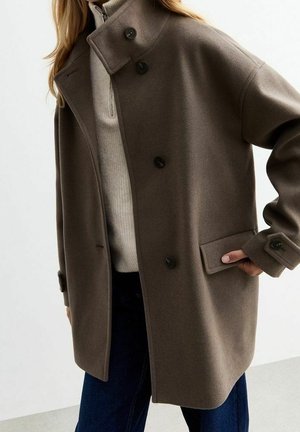 Žena v nadměrném taupe vlněném kabátu s černými knoflíky přes světle béžový žebrovaný svetřík se zipem a tmavě modré džíny.