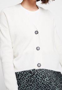 Cárdigan blanco de canalé con cuello en V, adornado con tres botones redondos oscuros, combinado con una falda verde con estampado de leopardo.