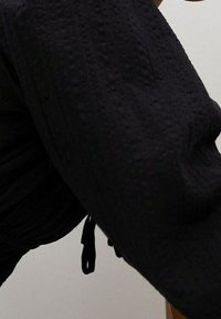 Schwarzer, strukturierter Stoff mit einem erhabenen Muster; hat eine lockere Passform und Raffung am Bund; seitliche Bänder sind sichtbar.